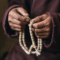 Weathered hands in maroon wool sleeves holding a yak bone barrel bead mala, the ivory beads banded with brown and grey