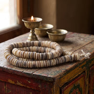 A yak bone mala coiled on a painted wooden cabinet beside a lit brass butter lamp and two small brass bowls by a window