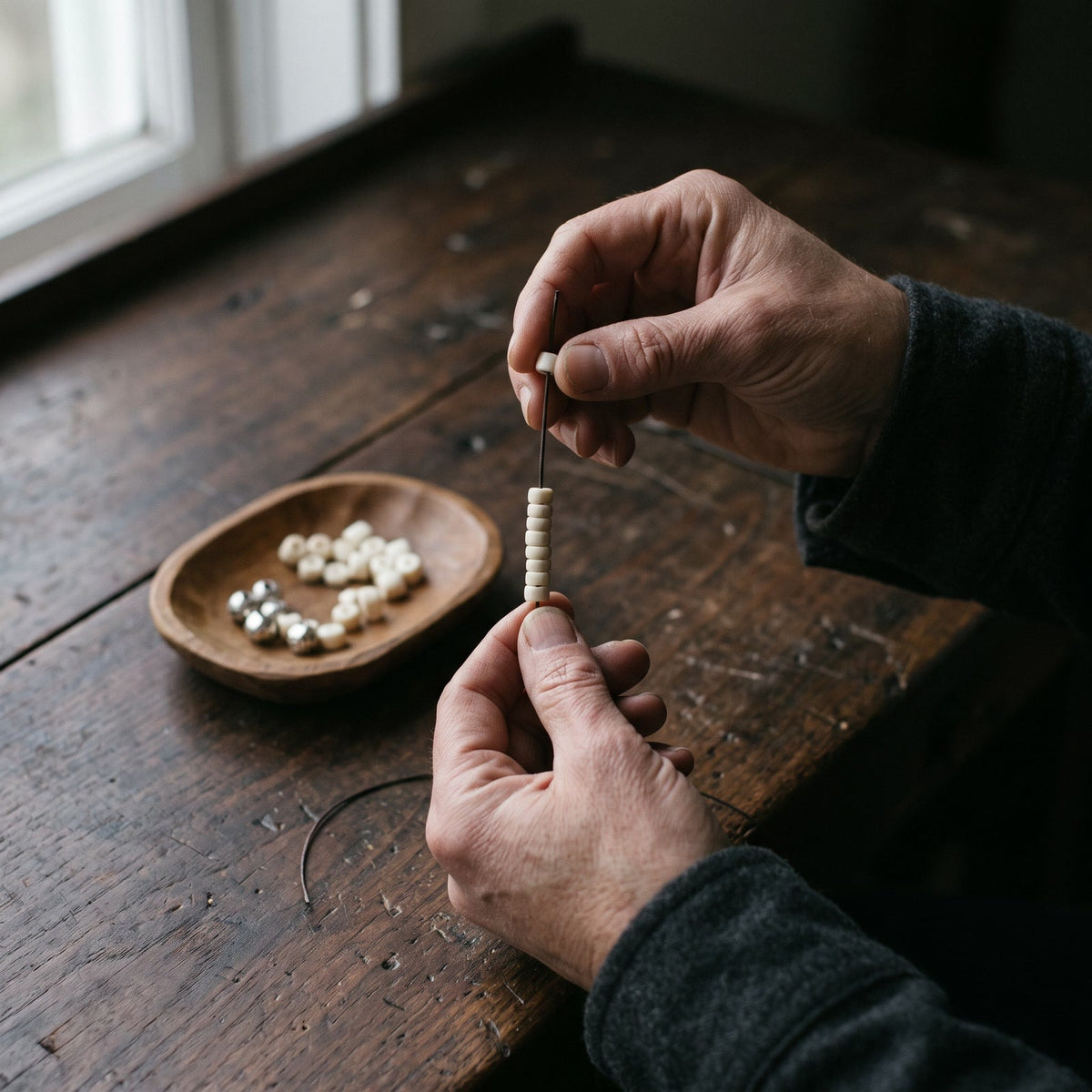 Hands threading bone beads onto a cord over a worn wooden workbench, a small wooden dish of loose beads nearby