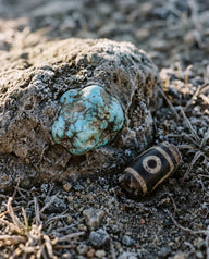 A raw turquoise nugget set in frost-rimmed host rock on stony ground, a worn striped bead in the soil beside it