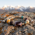 Beige braided cord bracelet with a tiger-tooth pattern dzi bead at its center on a lichen-covered boulder in low dawn light, snow peaks blurred behind