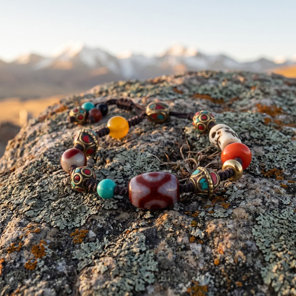 Dark brown braided cord bracelet with a three-eyed red dzi bead at its center, inlaid copper beads and a carved bone skull bead on a lichen-covered boulder in low dawn light, snow peaks blurred behind