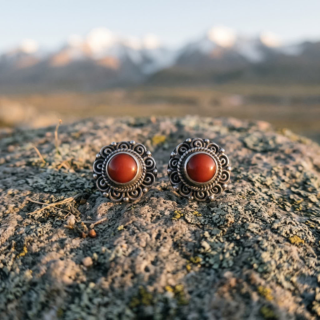 Pair of round silvery filigree stud earrings with red stones on a lichen-covered rock at dawn, snow peaks blurred behind