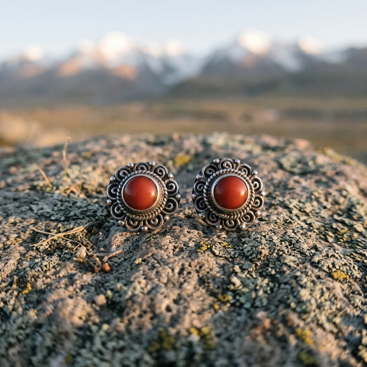 Pair of round silvery filigree stud earrings with red stones on a lichen-covered rock at dawn, snow peaks blurred behind