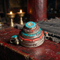 Tiered gau box pendant with a golden Om symbol on stabilized turquoise, red stones and silvery rope trim, on a red lacquered altar by butter lamps