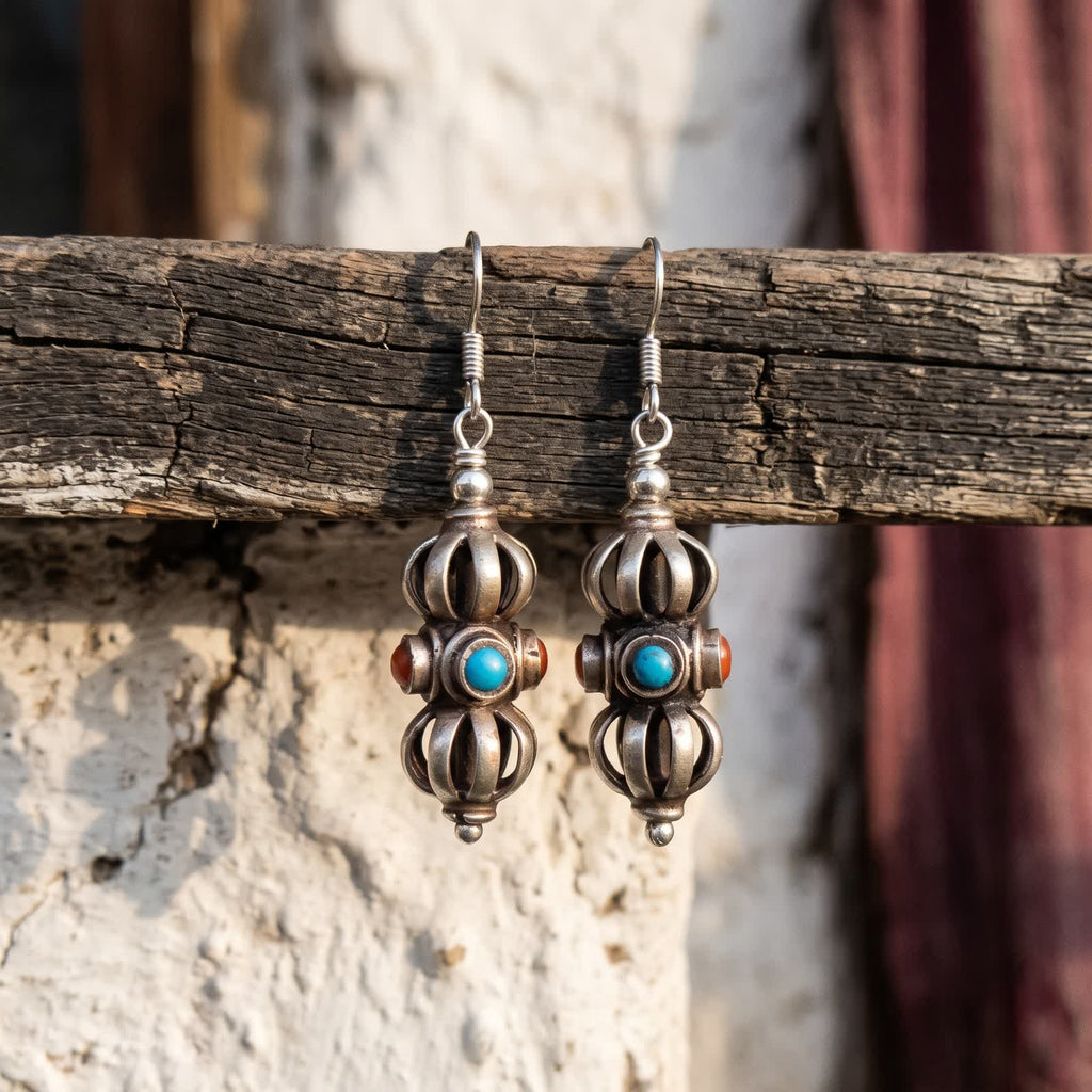 Pair of vajra drop earrings with red and blue stones, hanging from a weathered wooden rail against a whitewashed wall