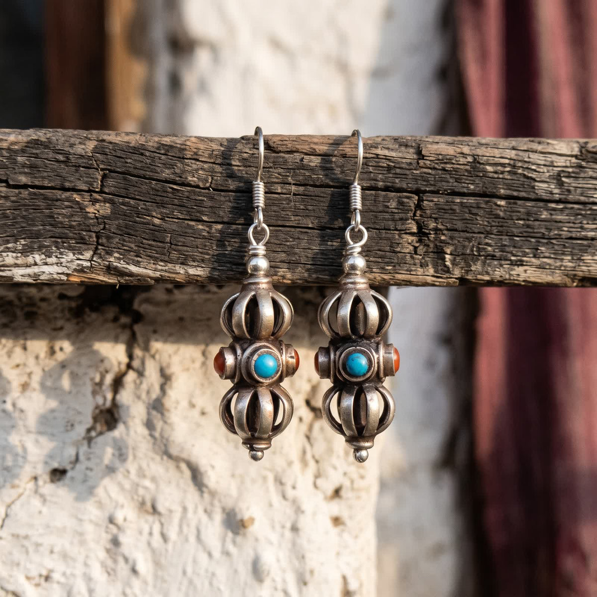 Pair of vajra drop earrings with red and blue stones, hanging from a weathered wooden rail against a whitewashed wall