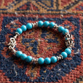 Blue turquoise bead bracelet with a silver vajra and small silver spacers on a worn red and indigo rug