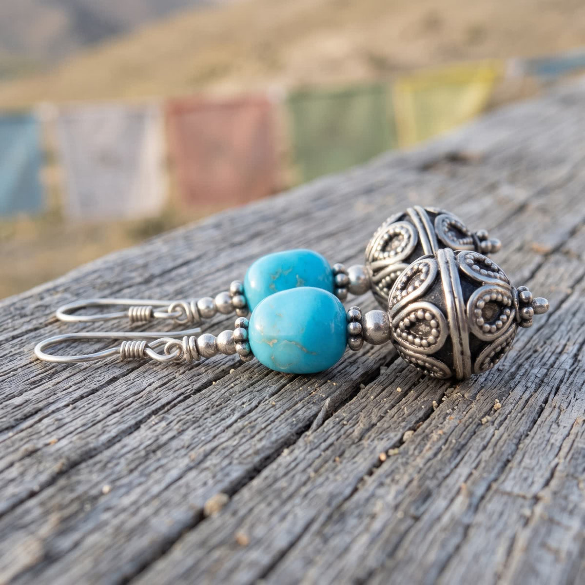 Pair of turquoise and silvery filigree ball drop earrings lying on weathered wood, prayer flags blurred behind