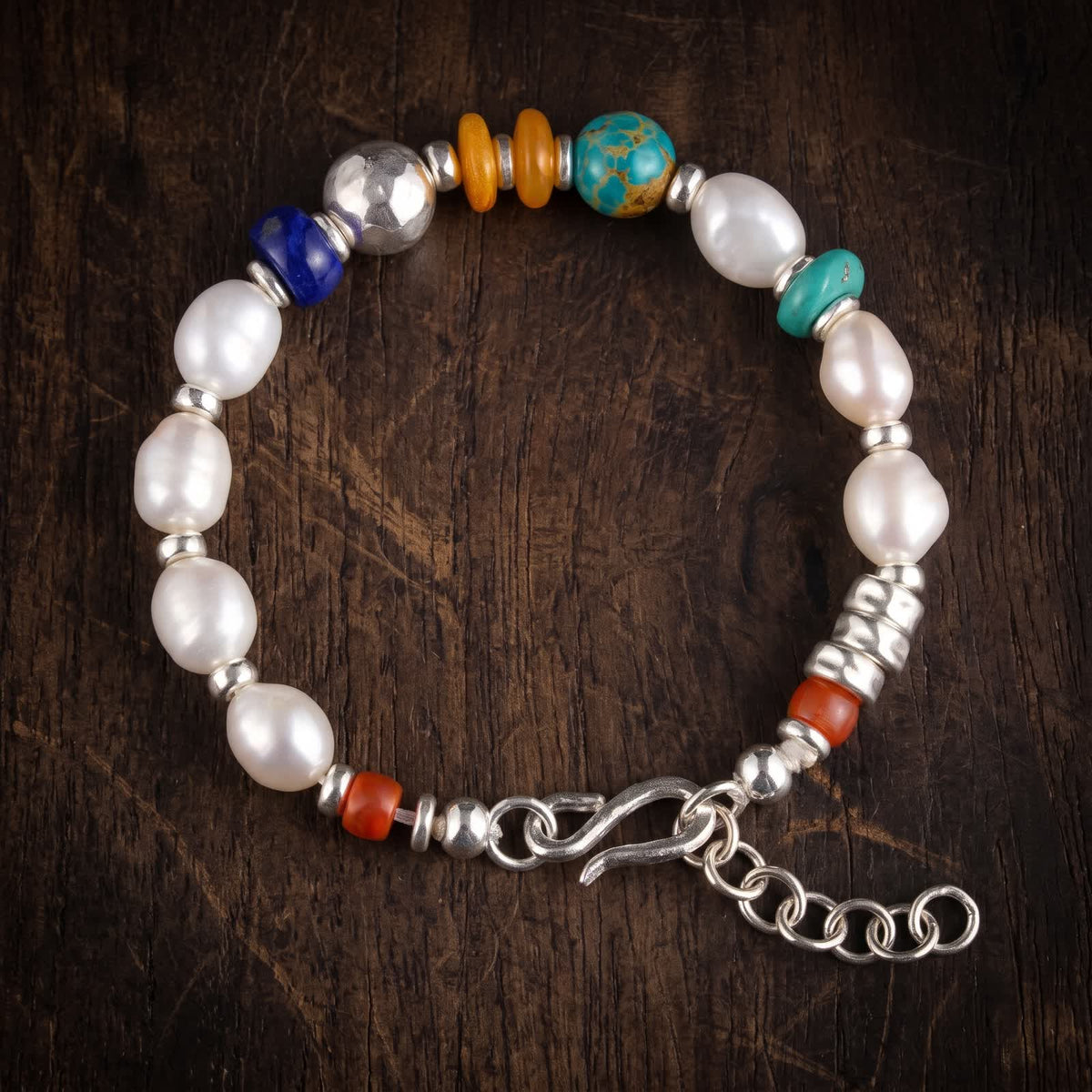 Pearl and mixed bead bracelet lying flat on dark wood: white pearls, a deep blue rondelle, a large hammered 925 sterling silver bead, two honey-orange discs and a blue-green bead, with a silver S-hook and extender chain