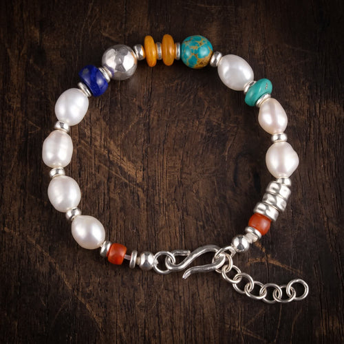 Pearl and mixed bead bracelet lying flat on dark wood: white pearls, a deep blue rondelle, a large hammered 925 sterling silver bead, two honey-orange discs and a blue-green bead, with a silver S-hook and extender chain
