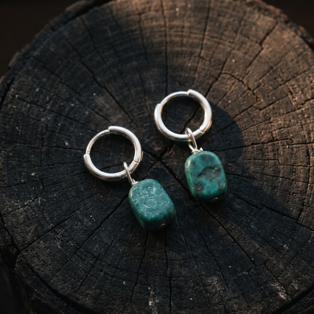 Pair of small hinged hoop earrings with green stone drops lying on a dark wooden stump in warm side light