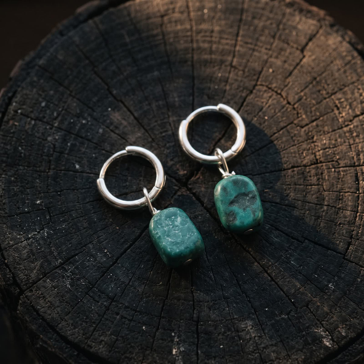 Pair of small hinged hoop earrings with green stone drops lying on a dark wooden stump in warm side light
