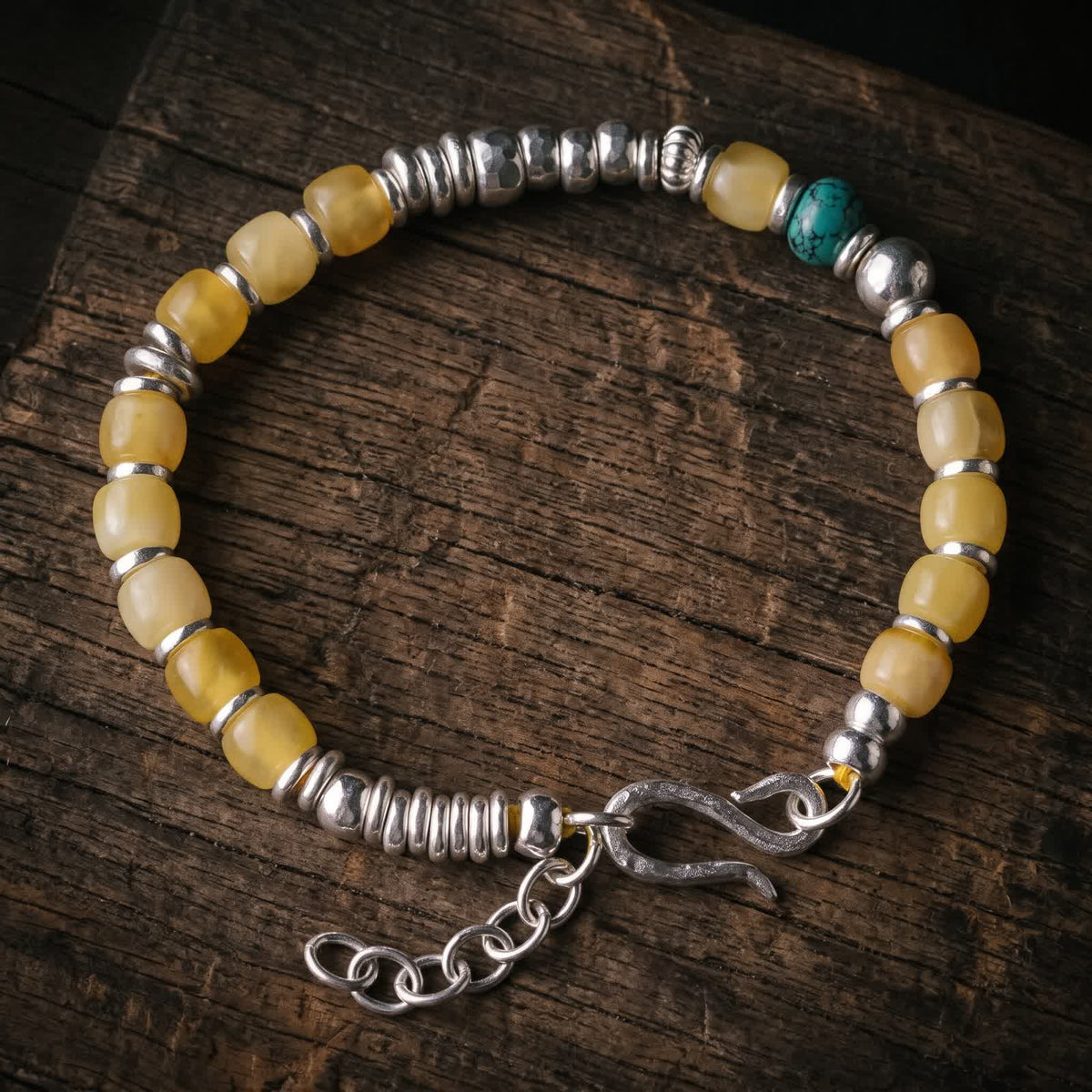 Honey-yellow bead bracelet with one turquoise bead, hammered 925 sterling silver beads and an S-hook clasp with extender chain, lying flat on dark wood