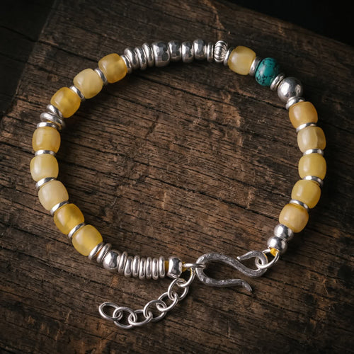 Honey-yellow bead bracelet with one turquoise bead, hammered 925 sterling silver beads and an S-hook clasp with extender chain, lying flat on dark wood