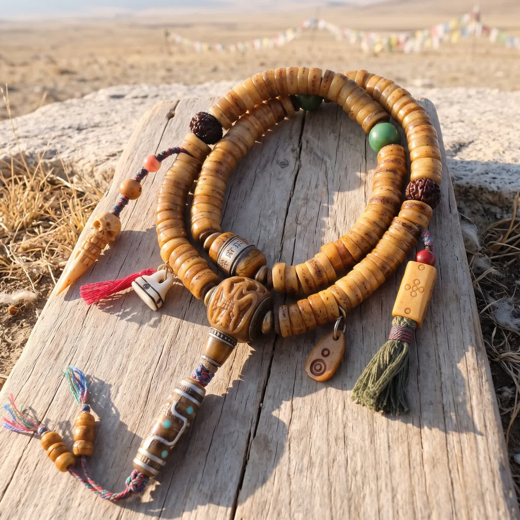 Honey yak bone disc mala coiled on sun-bleached wood in morning light, with carved bone guru bead, bone dzi-pattern bead, two counters and green and red tassels