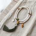 Pearl-white bead 108 mala laid in two loops on undyed linen and pale wood in soft window light, with a squared guru bead, green ribbed bead, dark green tassel, golden engraved white charm and orange drop charm