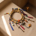 Striped bone disc 108 mala coiled on a pale plaster ledge in late-afternoon sun, hung with a skull guru bead, gau box pendant, bells, brass bead strings and blue, red and green tassels, a butter lamp glowing behind