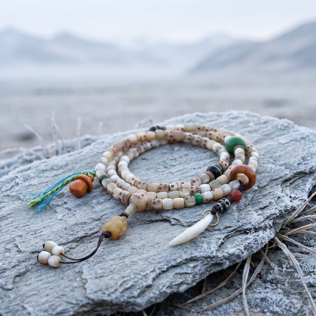Fish bone 108 mala with small carved skull faces coiled on frosted slate on an overcast morning, with a carved bone pendant, orange gourd charm and white tooth-shaped charm