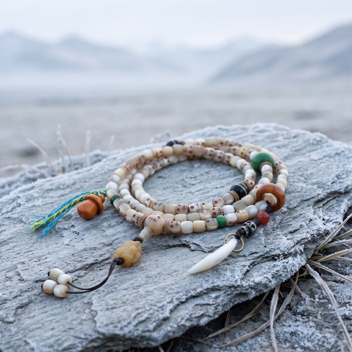 Fish bone 108 mala with small carved skull faces coiled on frosted slate on an overcast morning, with a carved bone pendant, orange gourd charm and white tooth-shaped charm