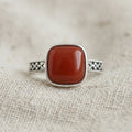 Close-up of an adjustable ring with a square red stone in a smooth bezel and an openwork band, resting on oatmeal linen