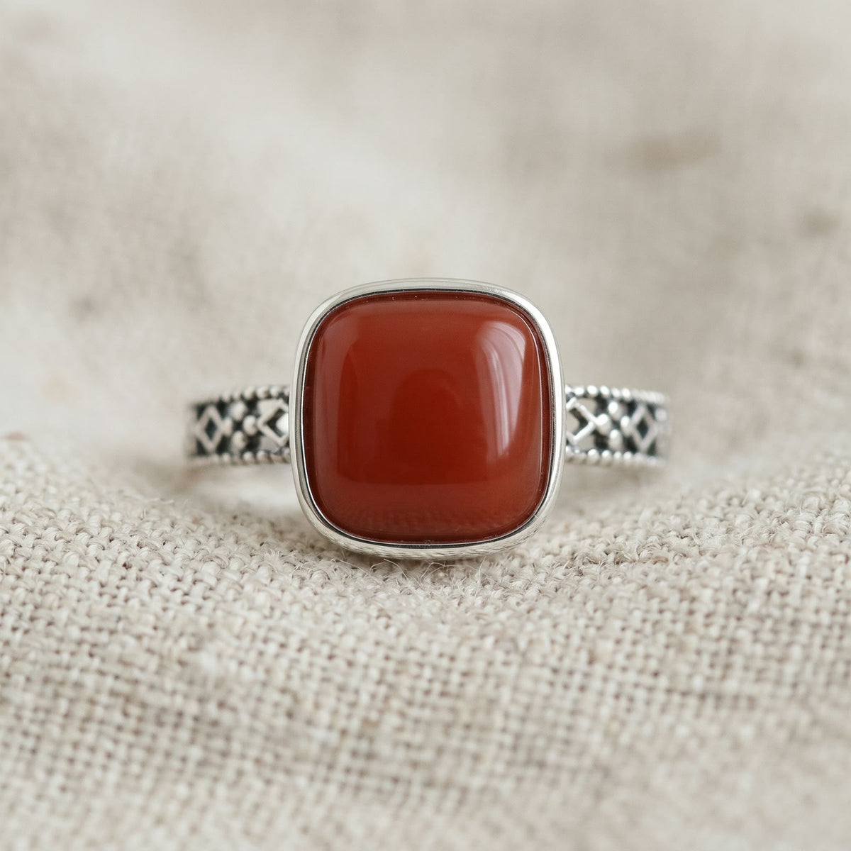Close-up of an adjustable ring with a square red stone in a smooth bezel and an openwork band, resting on oatmeal linen