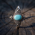Close-up of an adjustable ring with a small oval turquoise in a granulated, darkened setting with spiral scrolls and two fleur tips, on a dark wooden stump