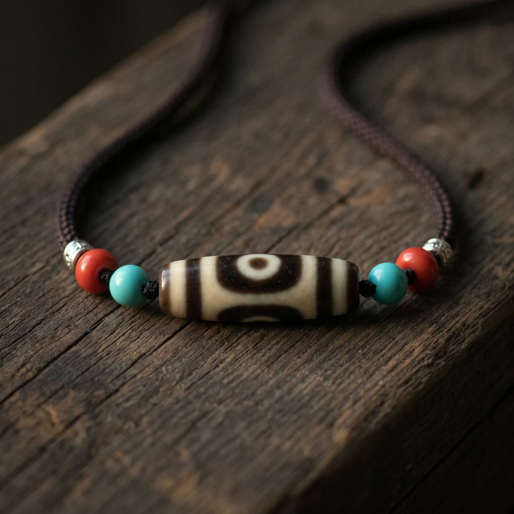 Two-eyed agate dzi bead necklace on a dark brown braided cord, flanked by blue-green beads, red beads and spacers, resting on an old weathered wood plank in soft warm side light