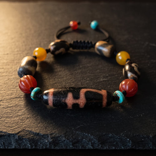 Red-vein vase-pattern agate dzi bead bracelet on black cord with four small agate dzi beads, red melon-carved beads, honey-yellow beads and blue-green discs, on dark slate in low warm side light