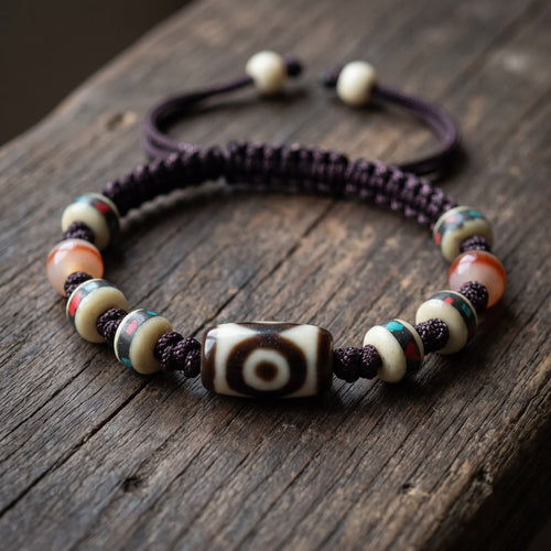 Eye-pattern agate dzi bead bracelet on a plum-brown braided cord, with cream-white inlaid disc beads and two red-band beads, resting on a weathered wood plank in soft warm side light