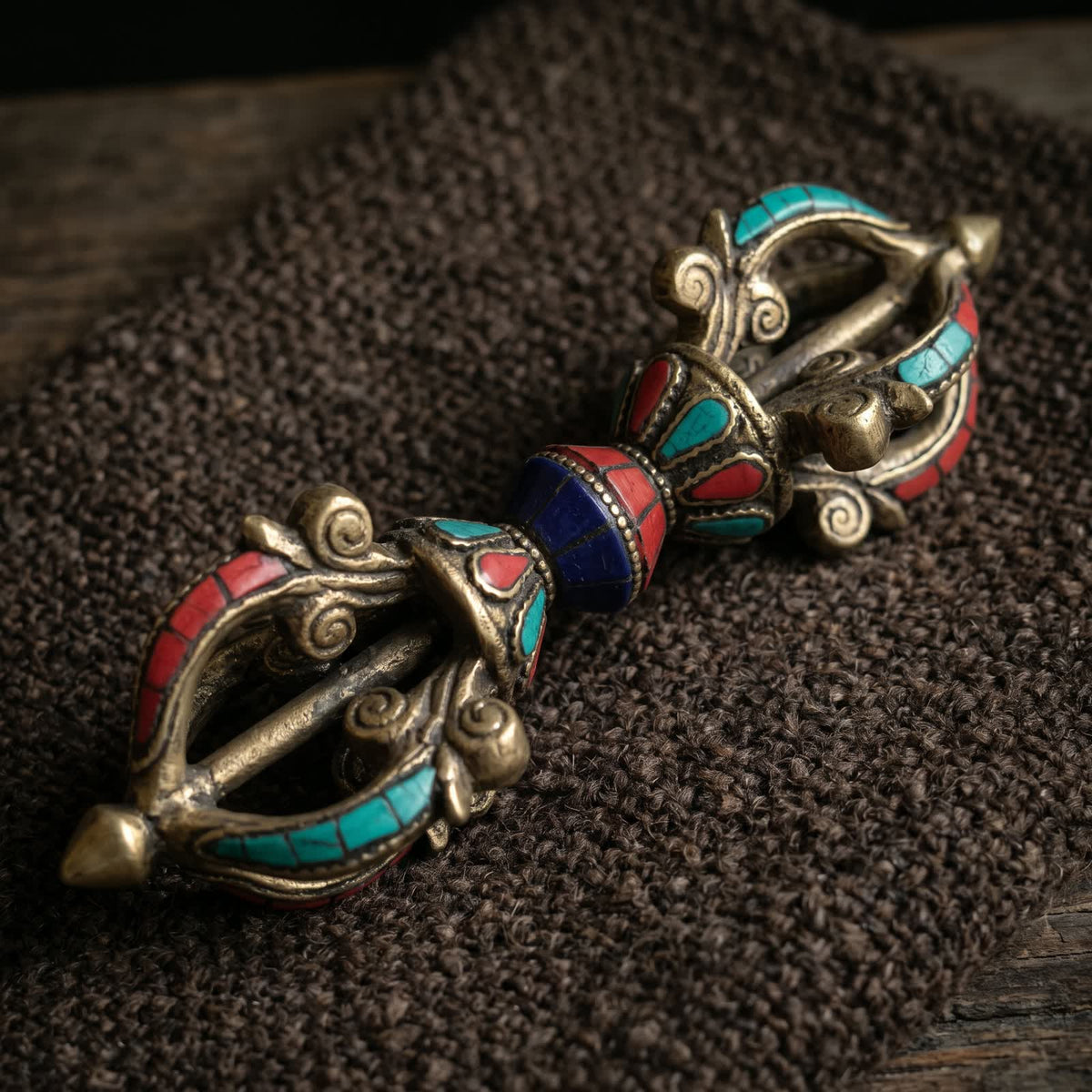 Vajra with red, blue-green and deep blue inlay, lying on dark brown handwoven wool cloth in soft warm side light