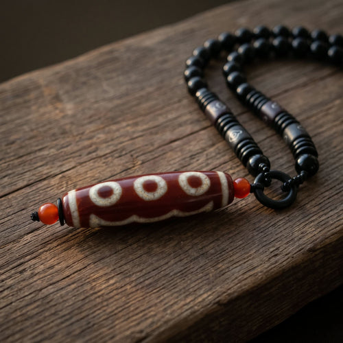 Red dzi bead pendant necklace on a strand of black beads, with small red beads at each end of the dzi and black carved barrel beads near the pendant, resting on a weathered wood plank in soft warm side light
