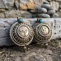 Two round gau box pendants side by side, one with Buddha eyes and one with the Om symbol, golden with copper-red centres, leaning on weathered wood by a fieldstone wall