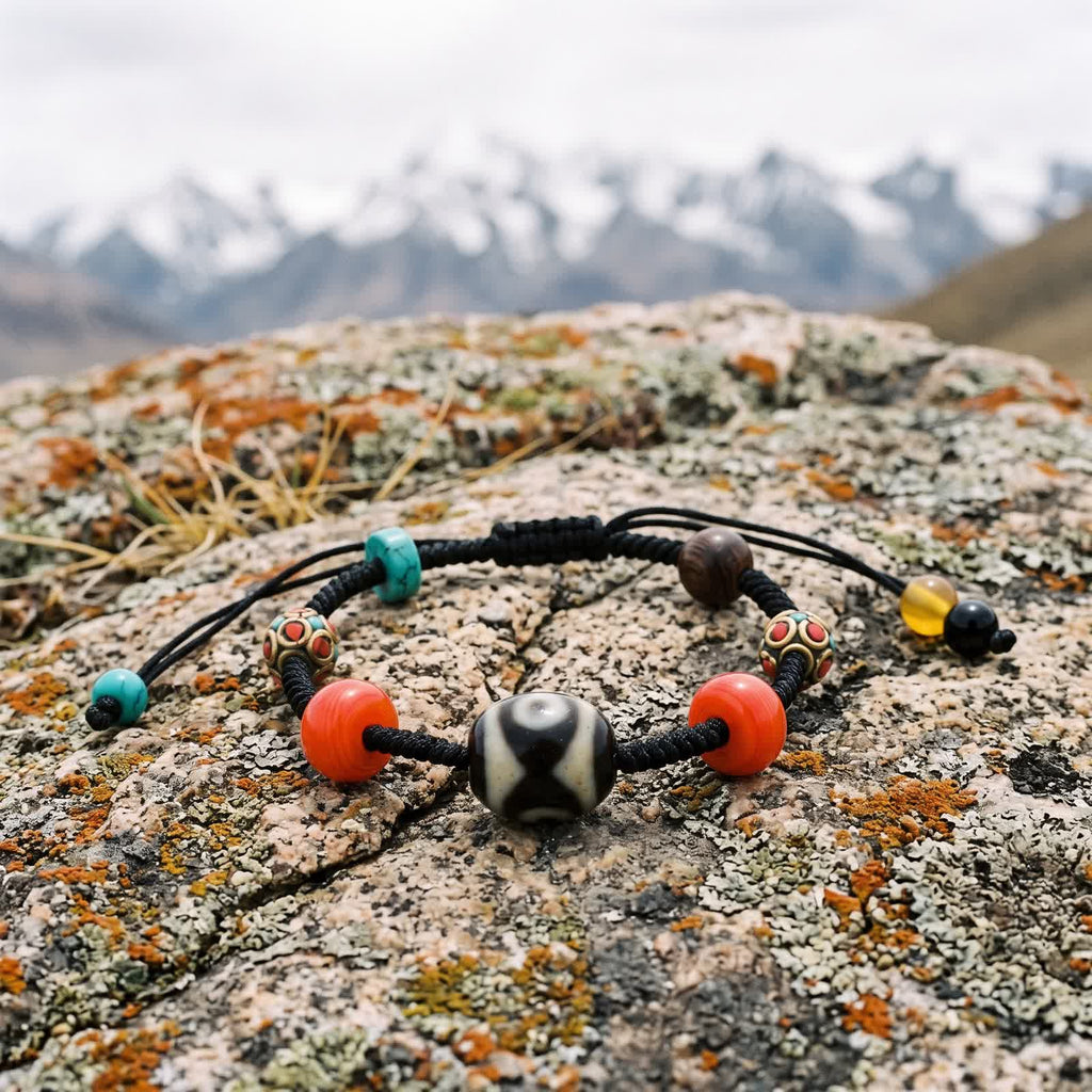 Black braided cord bracelet with a Dalo dzi bead between two red glass beads on a lichen-covered boulder, snow peaks blurred behind