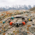 Black braided cord bracelet with a Dalo dzi bead between two red glass beads on a lichen-covered boulder, snow peaks blurred behind
