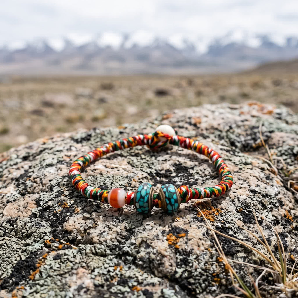 Five-color braided cord bracelet with a banded agate bead and two turquoise-chip inlay copper beads on a lichen-covered boulder, snow peaks blurred behind