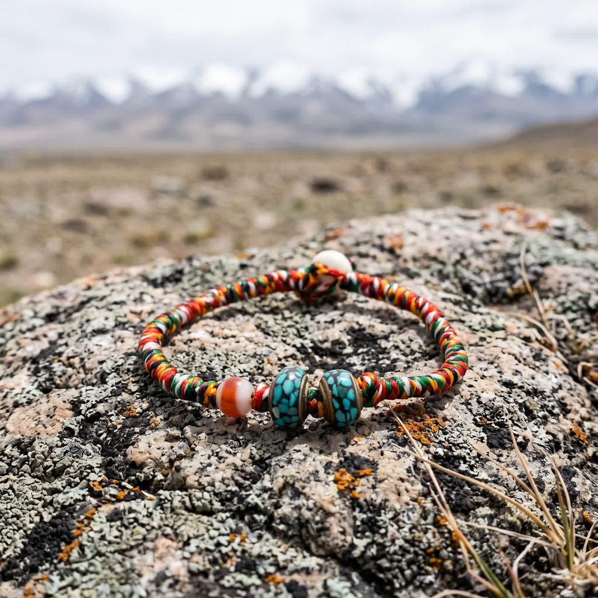 Five-color braided cord bracelet with a banded agate bead and two turquoise-chip inlay copper beads on a lichen-covered boulder, snow peaks blurred behind