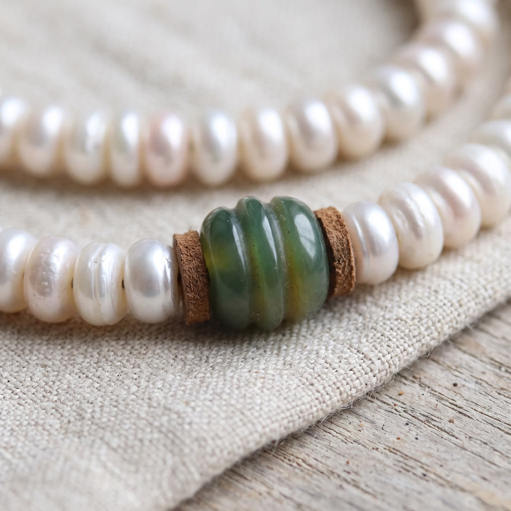 Close-up of lustrous flat pearl-white beads with small leather spacers on either side of a green ribbed bead, on undyed linen