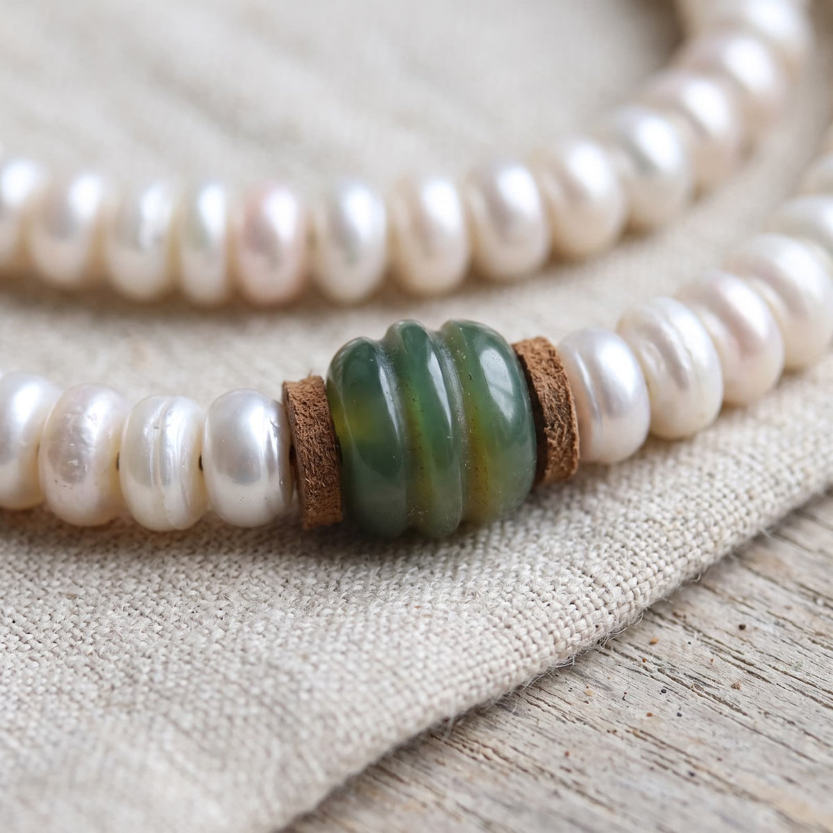Close-up of lustrous flat pearl-white beads with small leather spacers on either side of a green ribbed bead, on undyed linen
