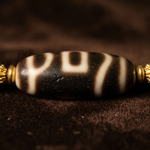 Macro close-up of the agate dzi bead, its ivory line pattern on deep brown, between small golden rondelles on dark brown velvet