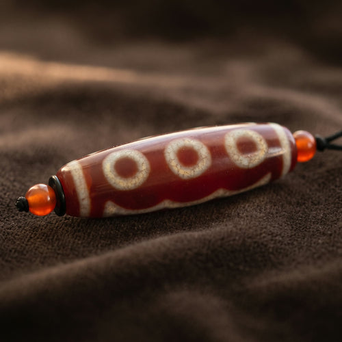 Macro close-up of the red dzi bead showing three cream-white circles above a wavy band, with a small red bead at each end, on dark brown velvet