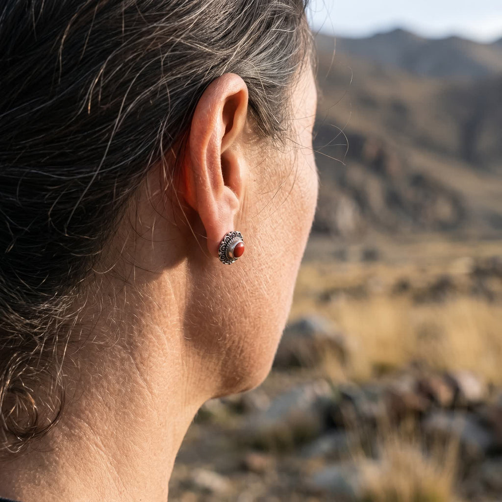 Red stone filigree stud earring on an earlobe, seen from behind with hair pulled back, sunlit hills blurred beyond