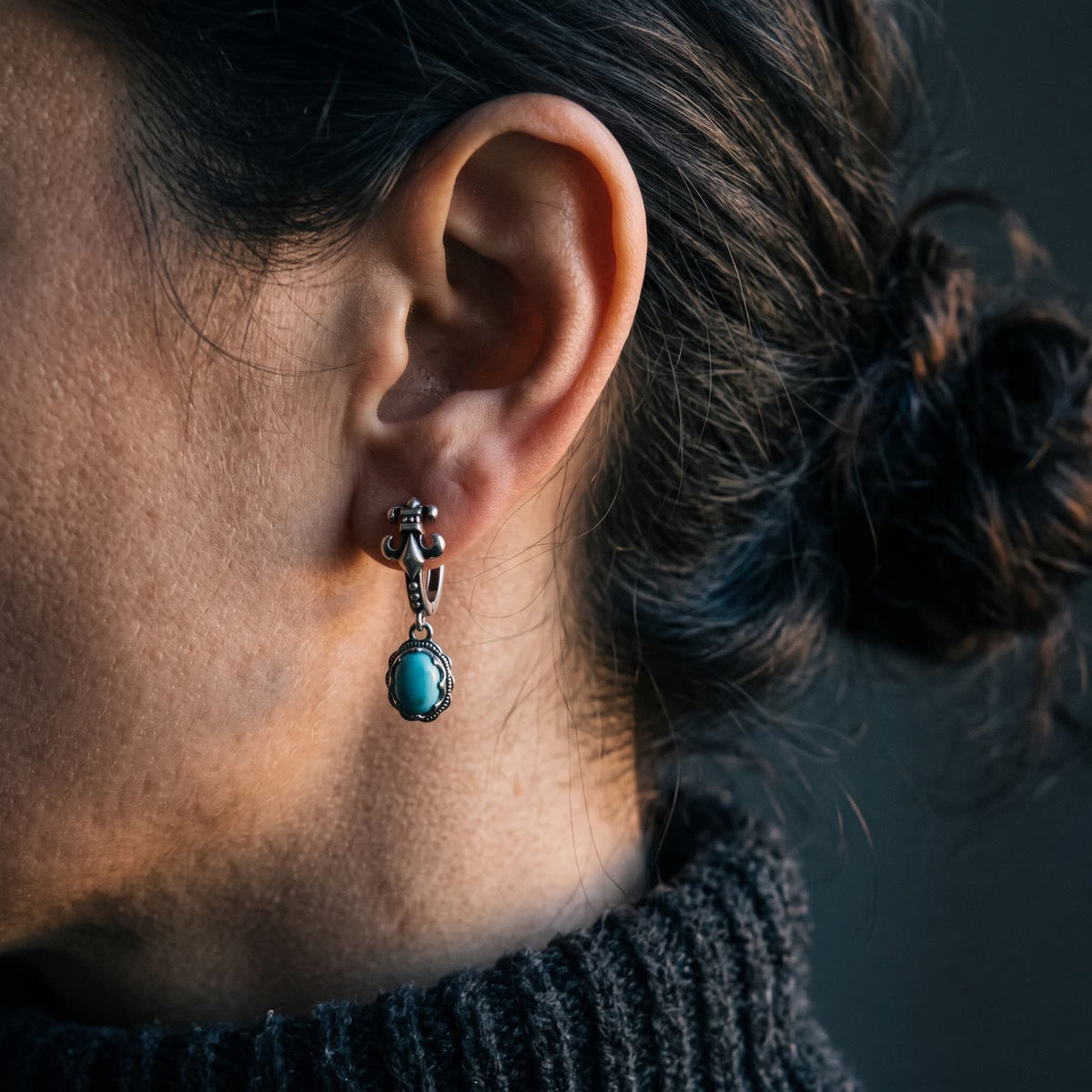 Fleur-de-lis hinged hoop earring with an oval turquoise drop worn on a woman's ear, dark background