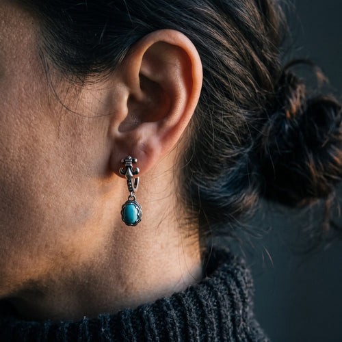 Fleur-de-lis hinged hoop earring with an oval turquoise drop worn on a woman's ear, dark background