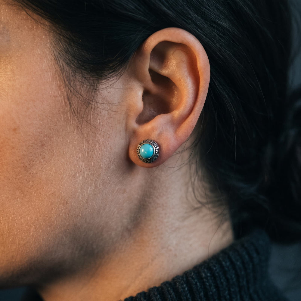 Round turquoise stud earring with a patterned sterling silver rim worn on a woman's earlobe, dark background