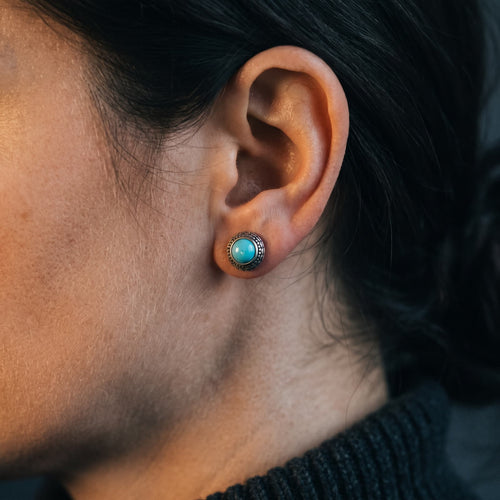 Round turquoise stud earring with a patterned sterling silver rim worn on a woman's earlobe, dark background