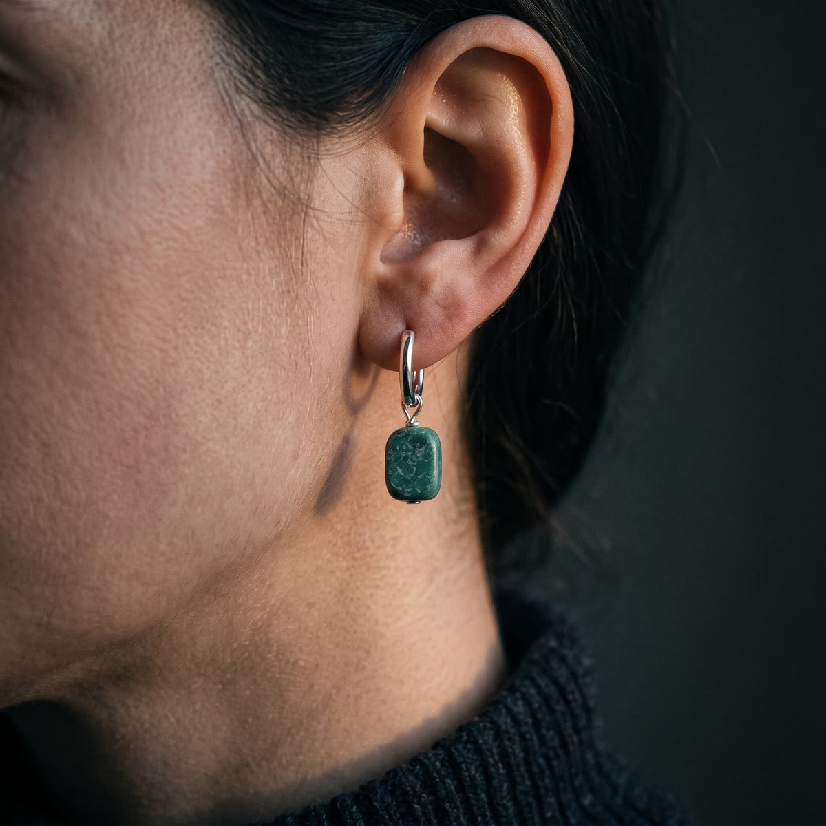 Small hinged hoop earring with a rectangular green stone drop worn on a woman's ear, dark background