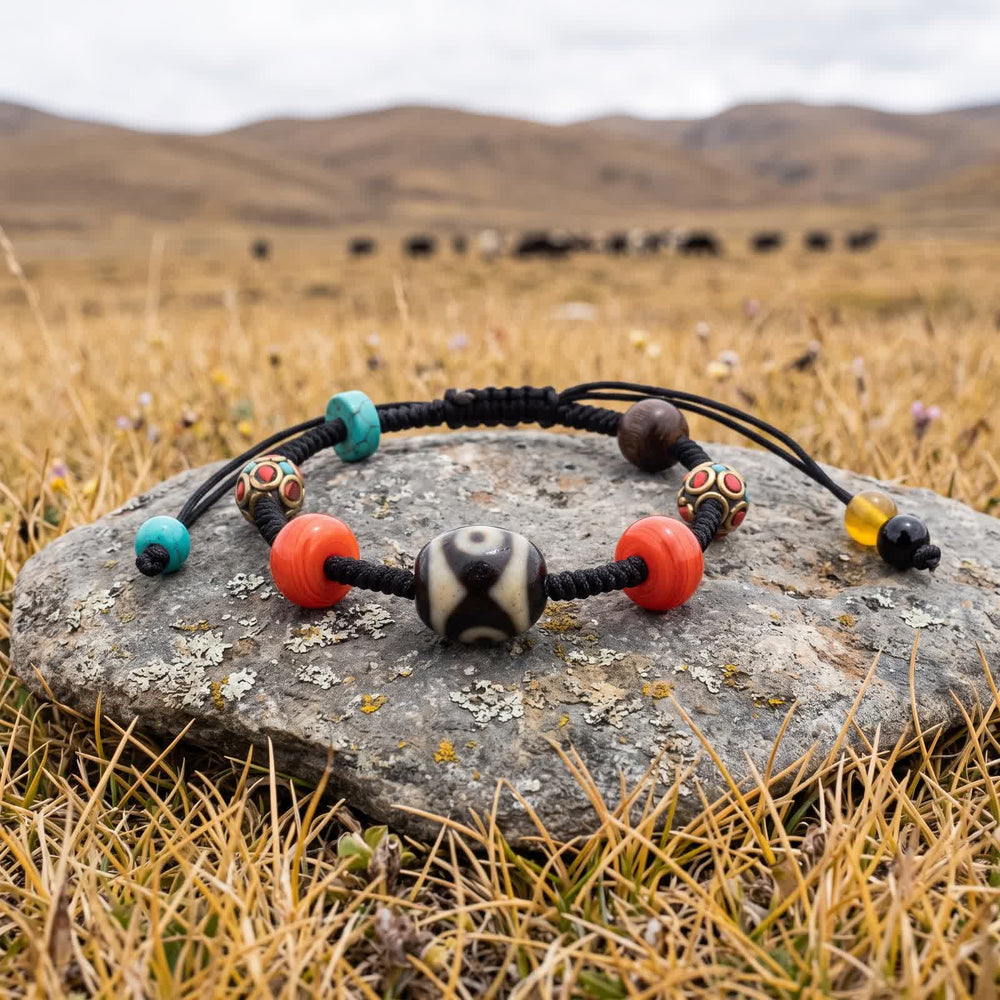 Dalo dzi cord bracelet on a flat lichen-spotted stone in dry plateau grass, a herd of yaks grazing in the distance