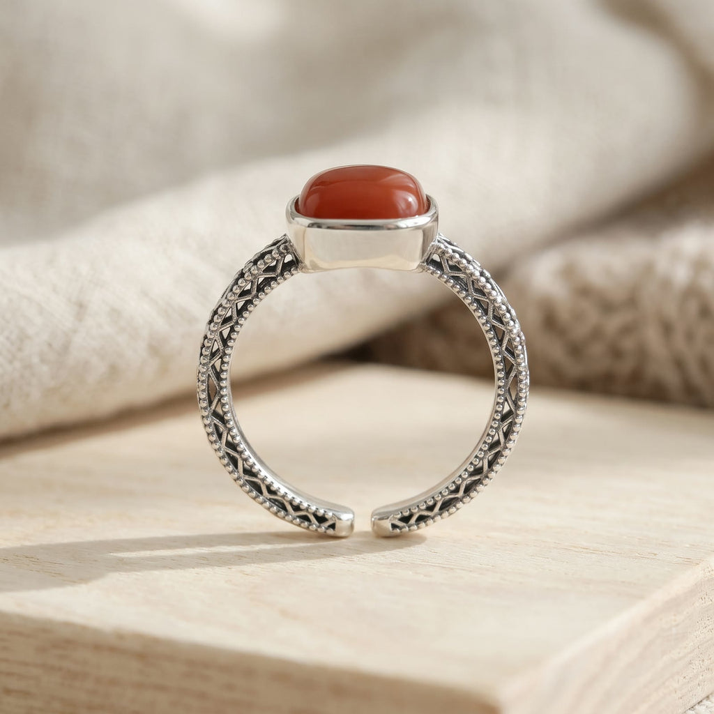Side view of the ring standing on pale wood, showing the openwork lattice band, beaded edges and the adjustable open band opposite the red stone