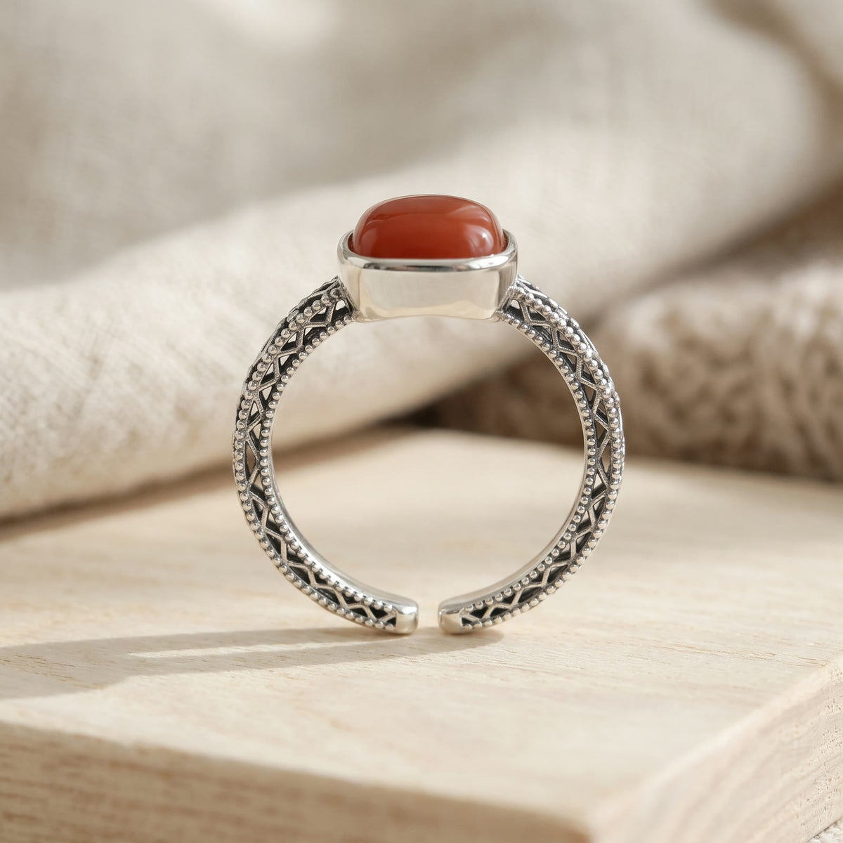Side view of the ring standing on pale wood, showing the openwork lattice band, beaded edges and the adjustable open band opposite the red stone